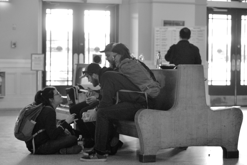 A group of young adults waits in a bus terminal, chatting.