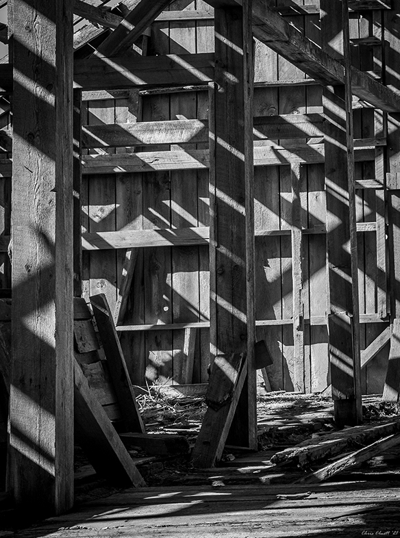 The inside of an old barn, taken at mid-day with sunlight streaming through a decayed roof and interacting with the barn’s structural skeleton.