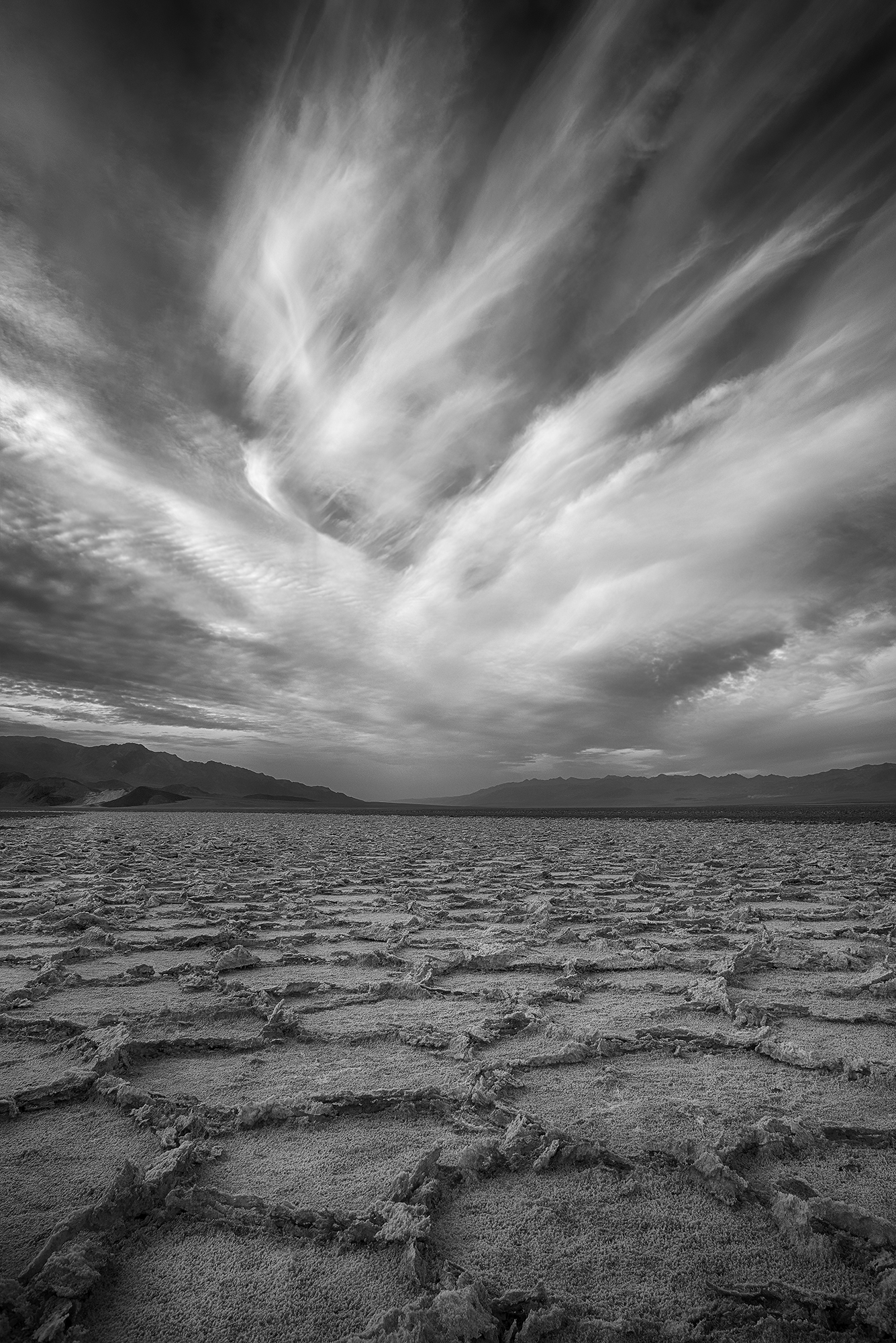 A black and white image showing a deserted desert and a cloudy sky. The ground is covered in cracks, and the horizon seems to stretch on forever.