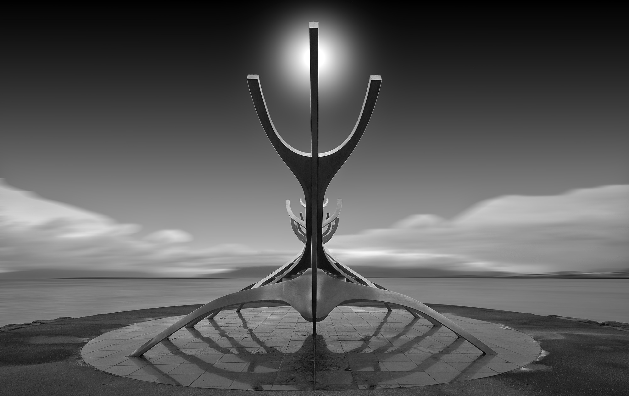 a black and white photo were a forked statue sits atop a circlar ceramic pad. The sun sets in the background over cloudy lake