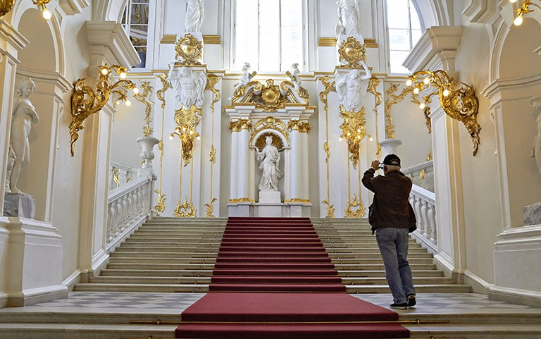 A shot of carpeted steps in the Hermitage museum leading to a statue with polls that have statue men holding post above their head containing gold filigree. the walls have gold light fixtures along the them.