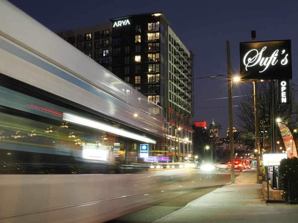 Long exposure night photograph of a city street with a motion-blurred train passing by. The illuminated Sufi’s restaurant sign, modern high-rise buildings, and streetlights create a dynamic urban scene. (25mm, f/9, 1/1.6 sec, ISO 1250, natural, no noise reduction)
