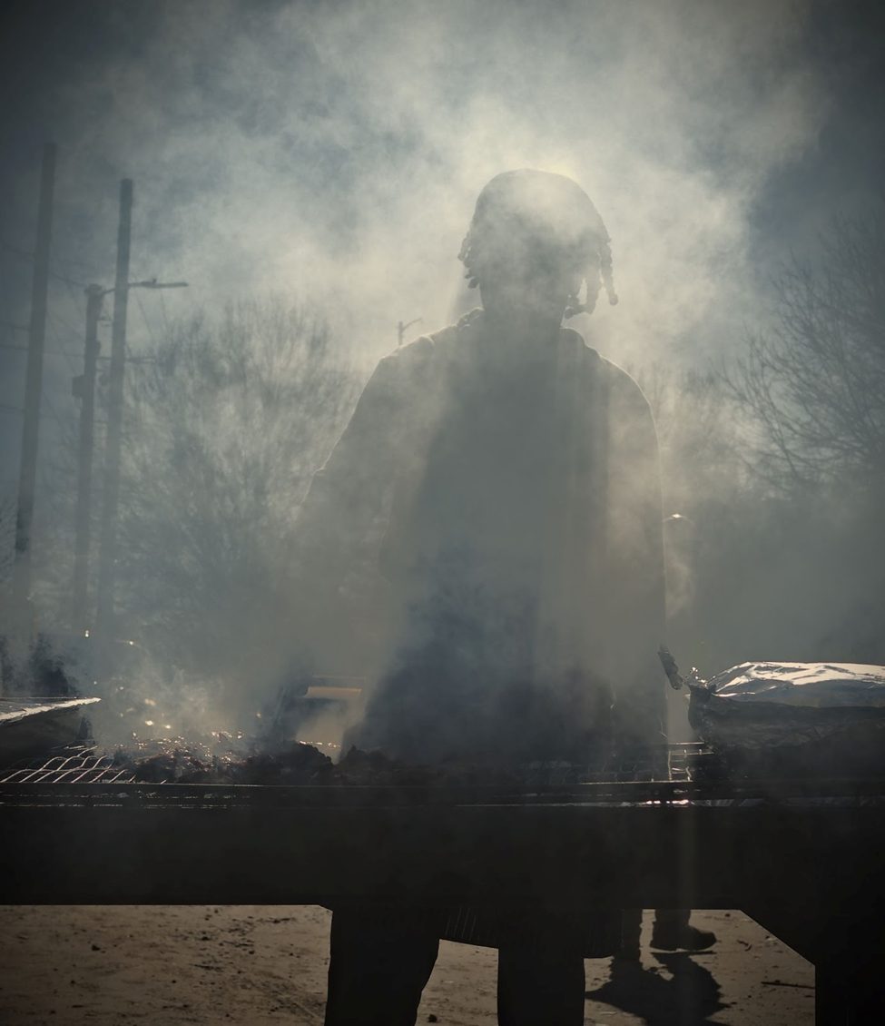 Silhouetted street food vendor grilling over smoky barbecue, backlit by natural sunlight. Atmospheric outdoor cooking scene. (25mm, ISO 100, f/5, 1/2000s)