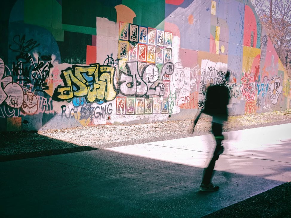 A dynamic street photography shot captured with the OM SYSTEM OM-3, featuring a blurred silhouette of a passerby against a graffiti-covered wall. The interplay of motion blur, urban textures, and vibrant street art showcases the energy and creativity of city life