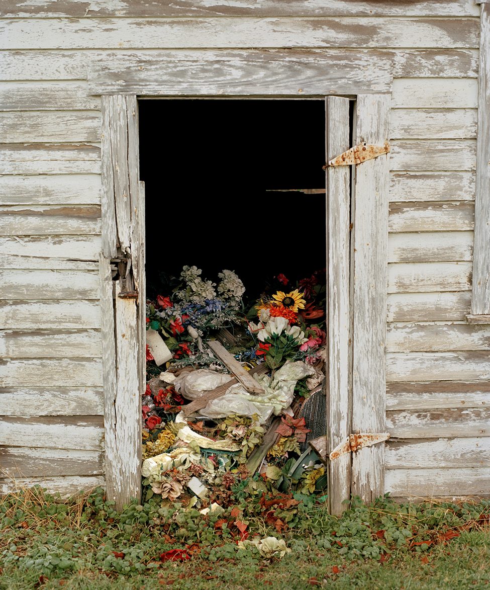 A decaying wooden shed with peeling white paint, overflowing with discarded artificial flowers and funeral decorations. Captured by photographer Dana Stirling for her analog photography book 'Why Am I Sad,' this image reflects themes of memory, abandonment, and emotional loss.