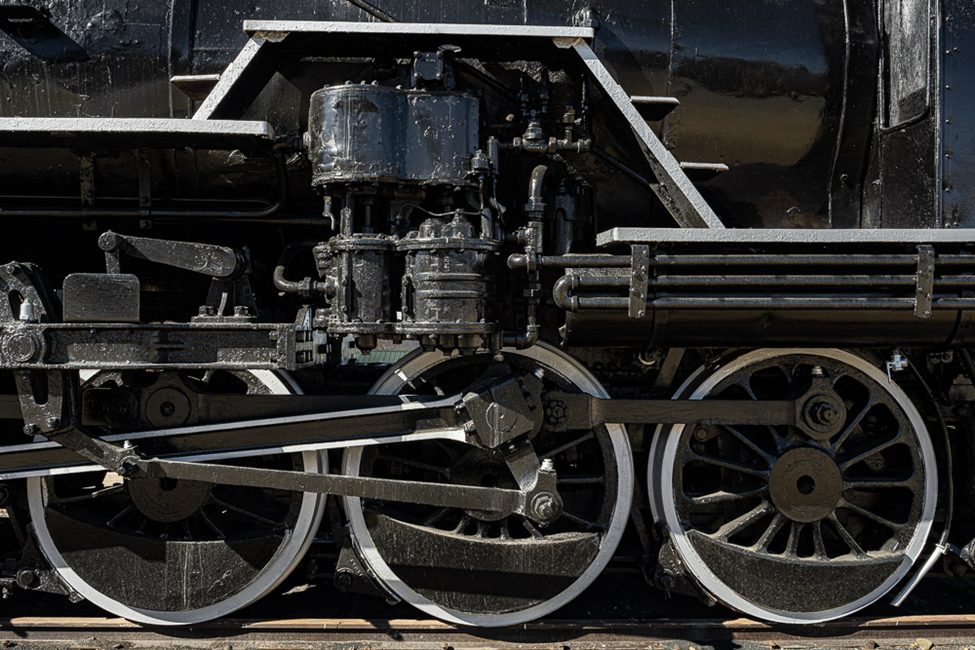 A close-up view of the intricate mechanical components and large metal wheels of a vintage steam locomotive. The textures and details of the industrial design highlight the beauty of classic engineering, perfect for capturing unique perspectives in photography