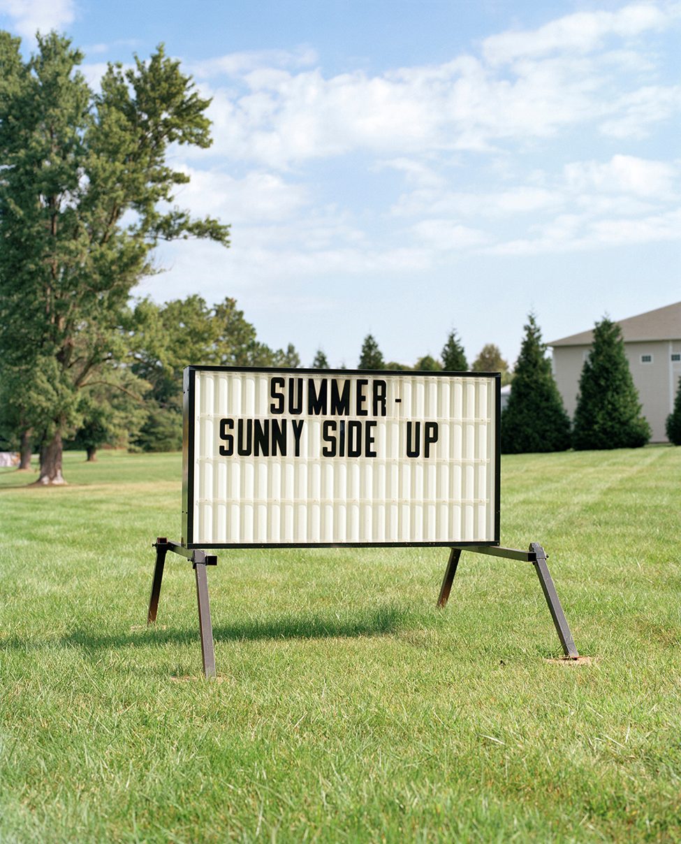 A vintage roadside sign reading 'SUMMER - SUNNY SIDE UP,' photographed by Dana Stirling as part of her analog photography book 'Why Am I Sad,' capturing subtle emotional contradictions and the search for beauty in everyday scenes