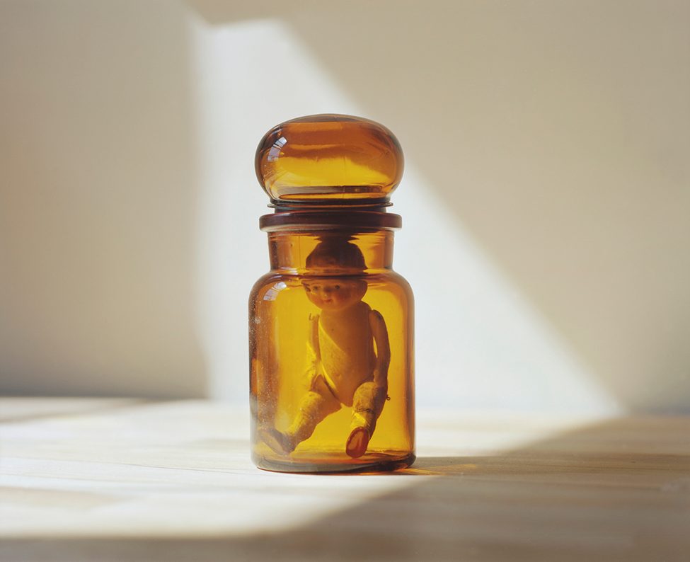 A vintage porcelain doll trapped inside an amber glass jar, captured in soft natural light by photographer Dana Stirling for her analog photography book 'Why Am I Sad,' reflecting themes of nostalgia and emotional vulnerability.