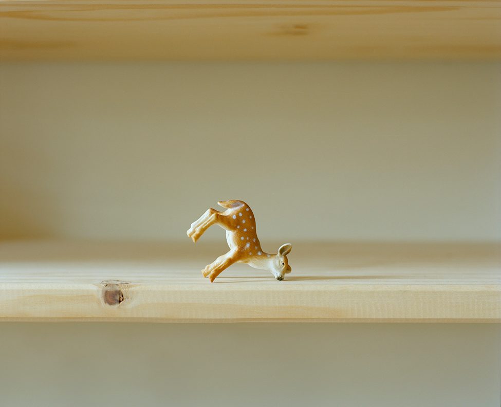 A delicate porcelain figurine of a fawn balancing on its head, photographed by Dana Stirling for her analog photography book 'Why Am I Sad.' The minimalistic composition and soft natural light evoke themes of fragility and emotional imbalance.