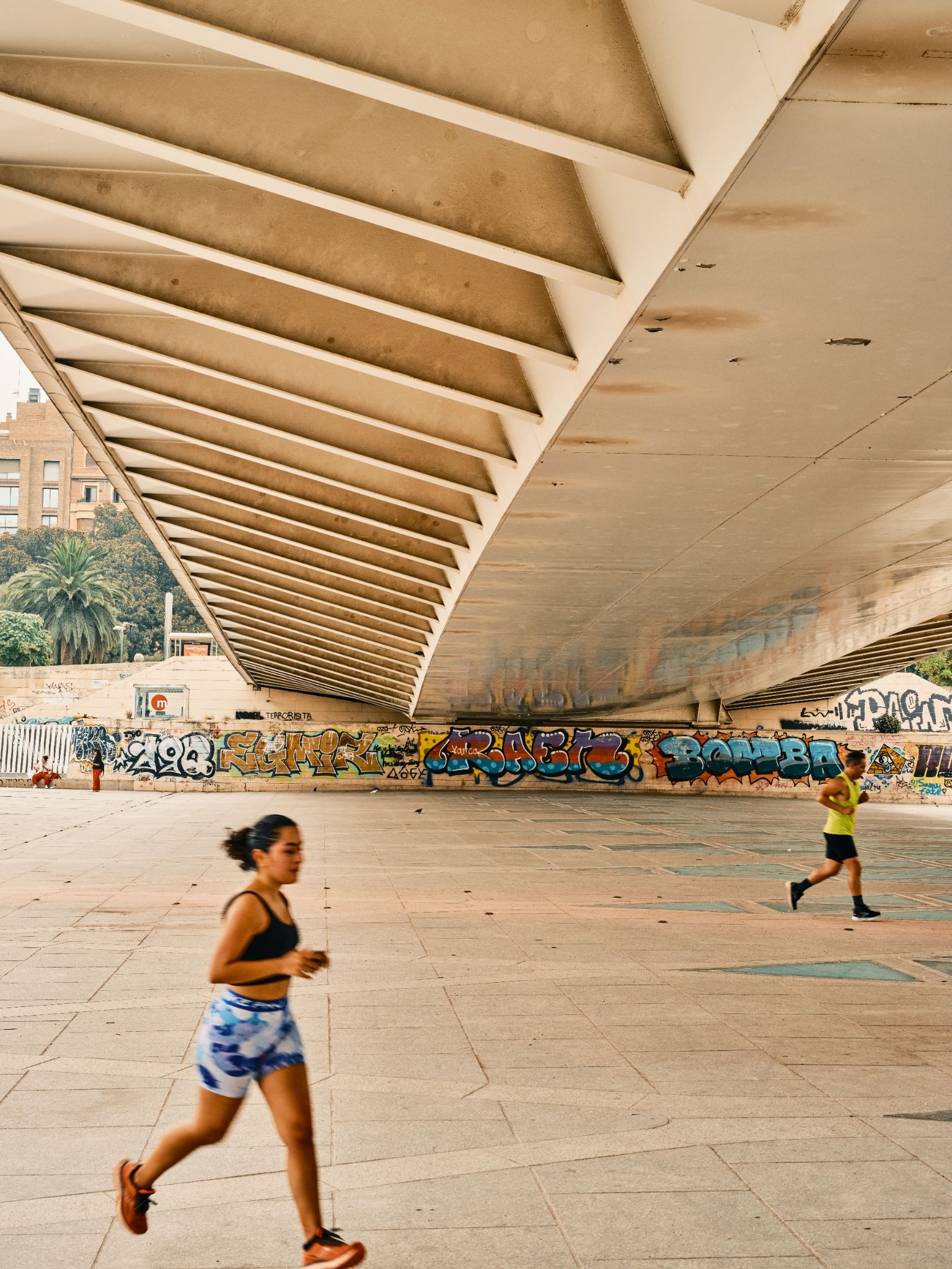 Joggers pass beneath a modern bridge covered with graffiti.