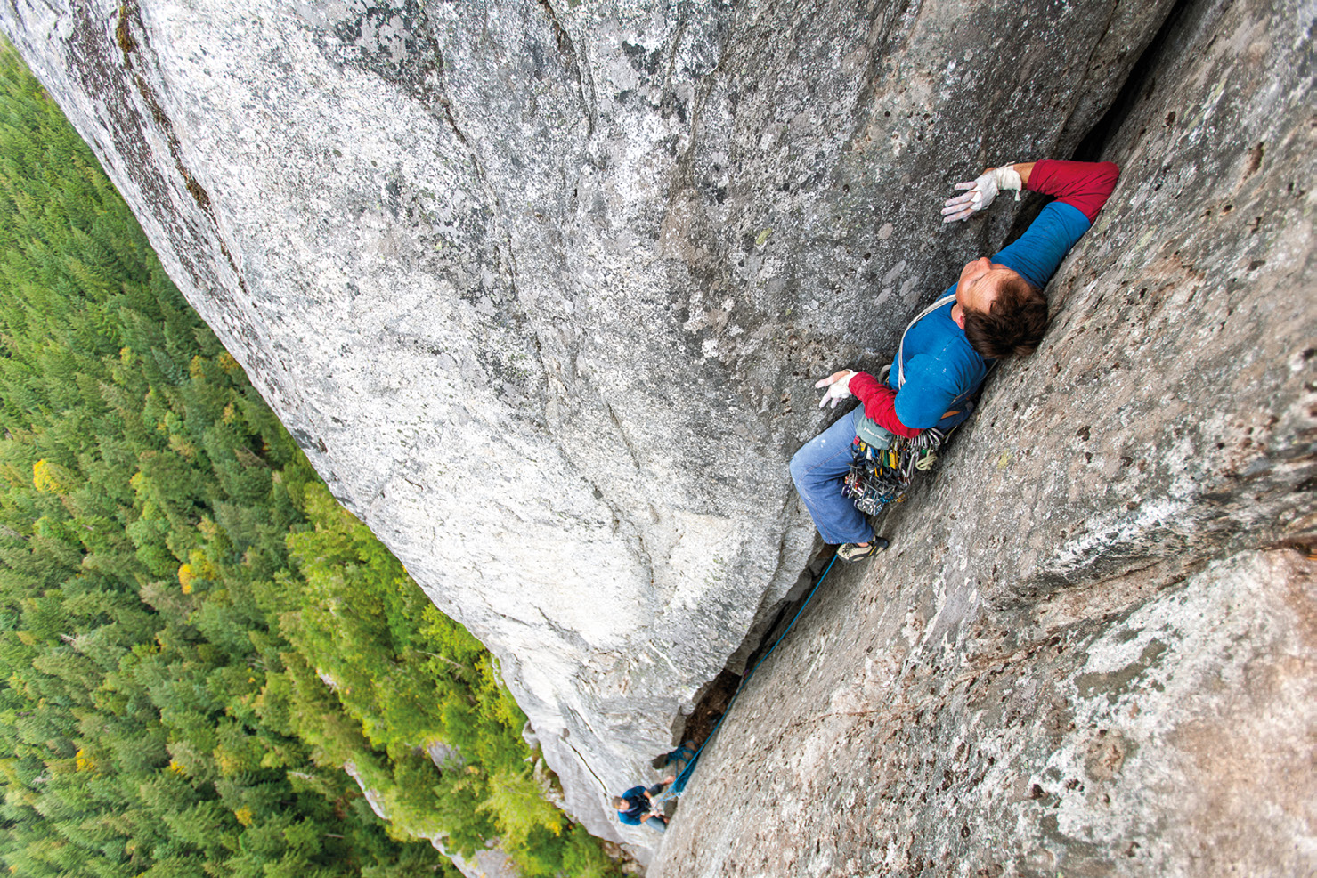 A climber ascending a steep granite rock face above a forested landscape.