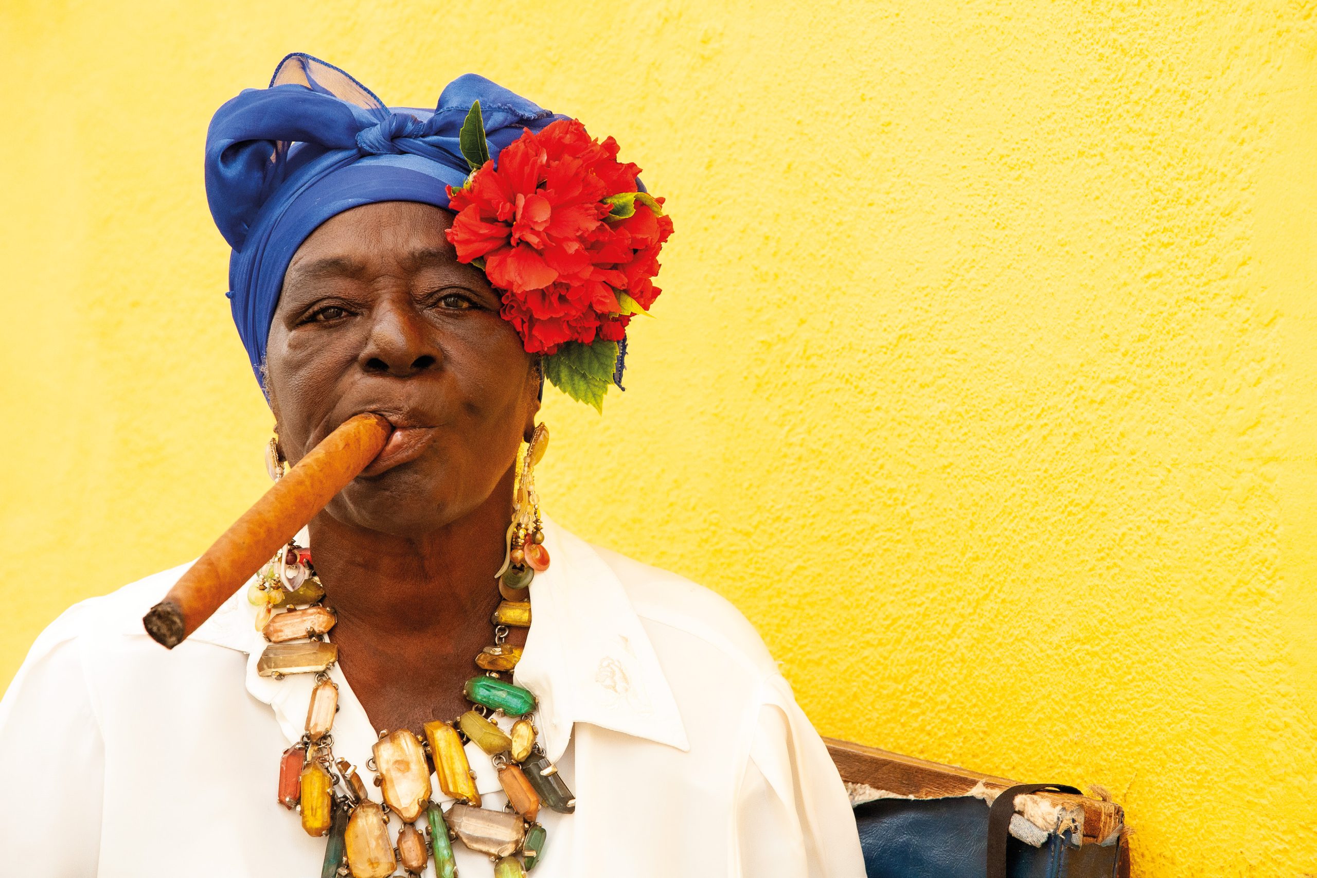A woman wearing a blue headwrap and red flower, smoking a cigar against a bright yellow wall.