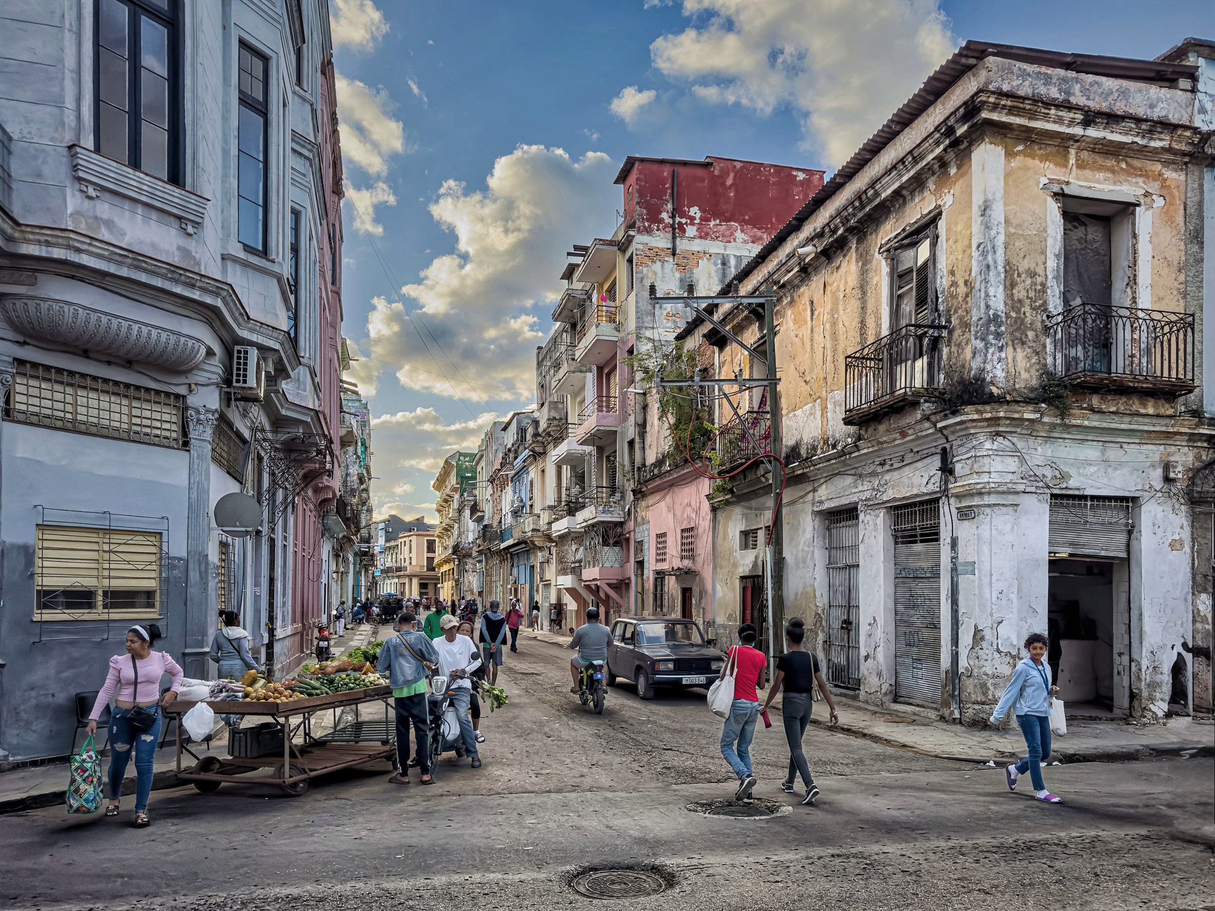 Street scene in Havana, Cuba, with colorful old buildings, pedestrians, and a vendor selling produce from a cart.