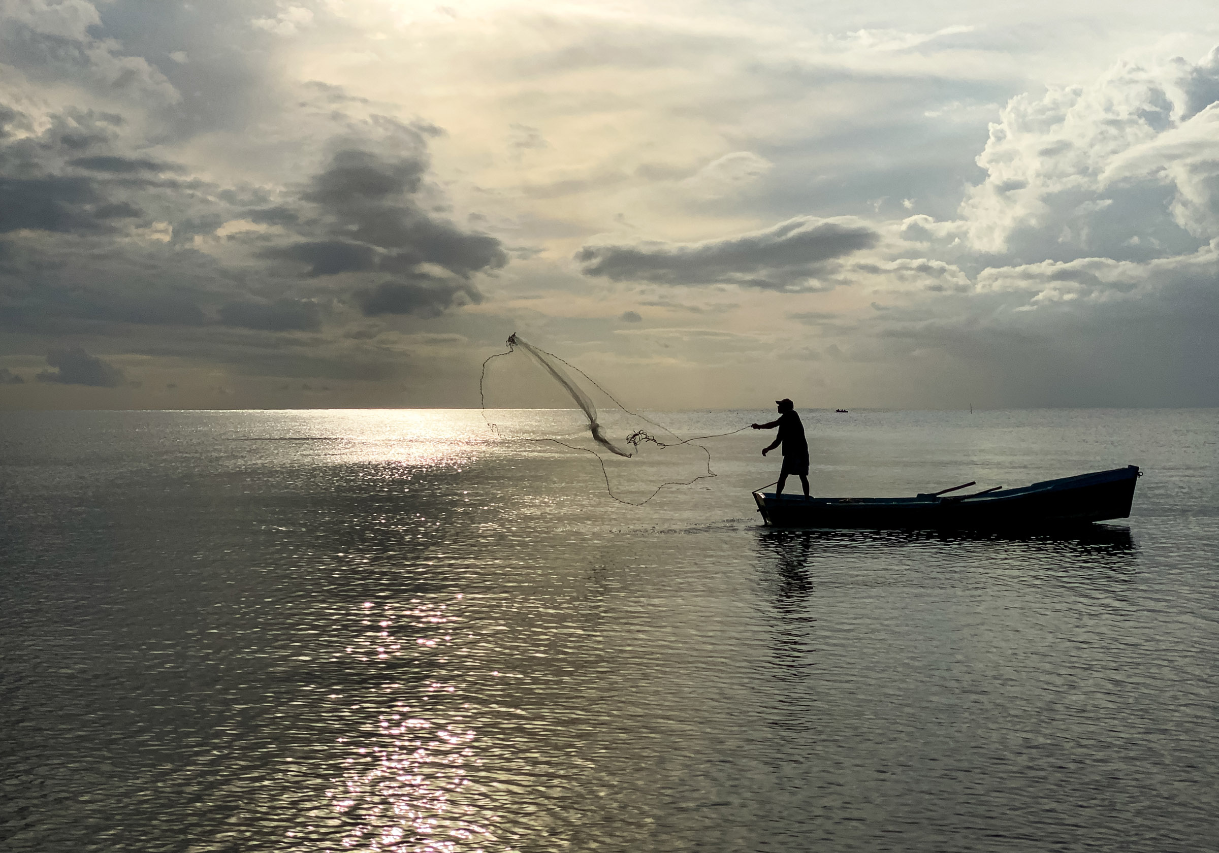 Fisherman casting a net from a small boat on the water at sunset.