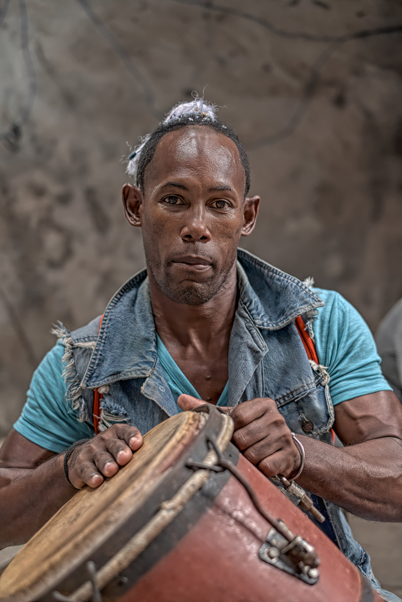 Cuban man wearing a turquoise shirt and denim vest, holding a wooden object.