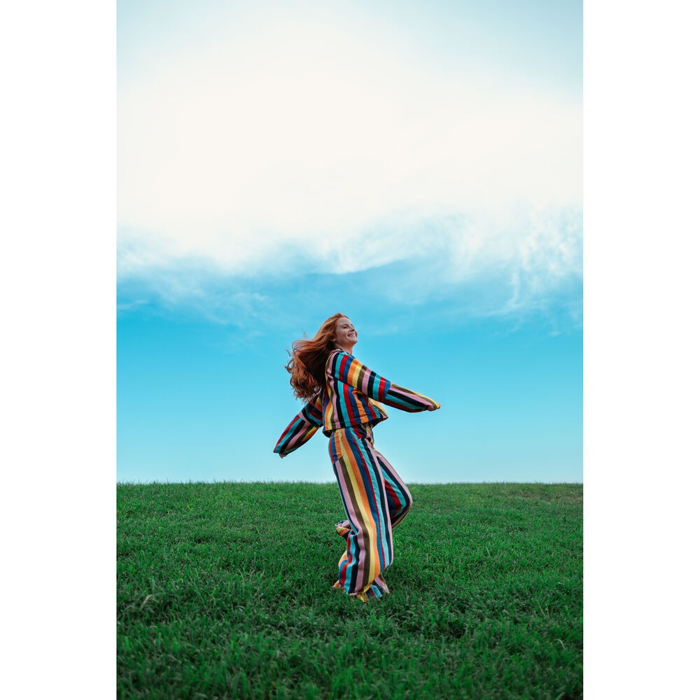 Woman wearing a colorful striped outfit standing in a grassy field under a blue sky.