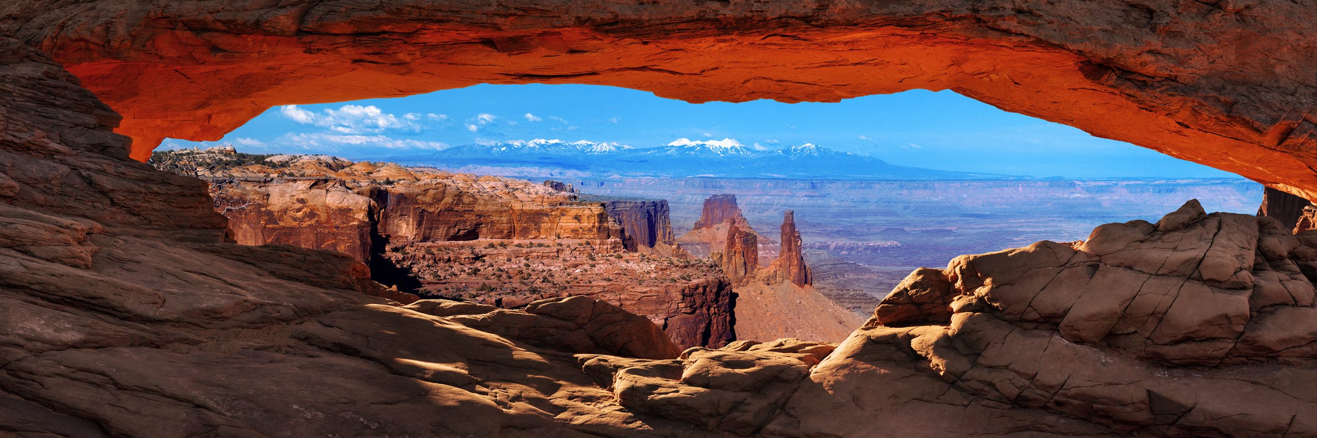 Mesa Arch framing distant canyon spires and snow-capped mountains beneath a blue sky