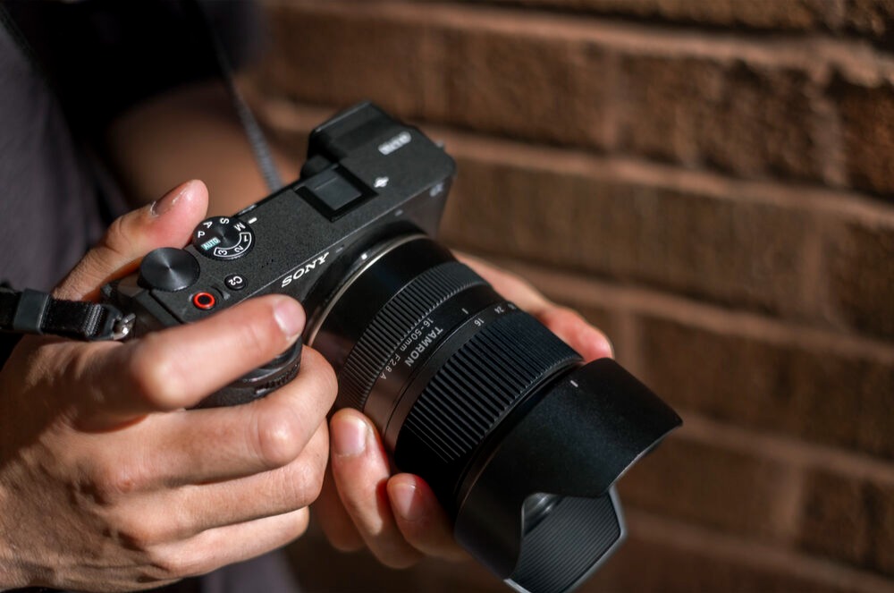 Sony mirrorless camera fitted with a Tamron 16-50mm f/2.8 zoom lens, held in a photographer's hands.