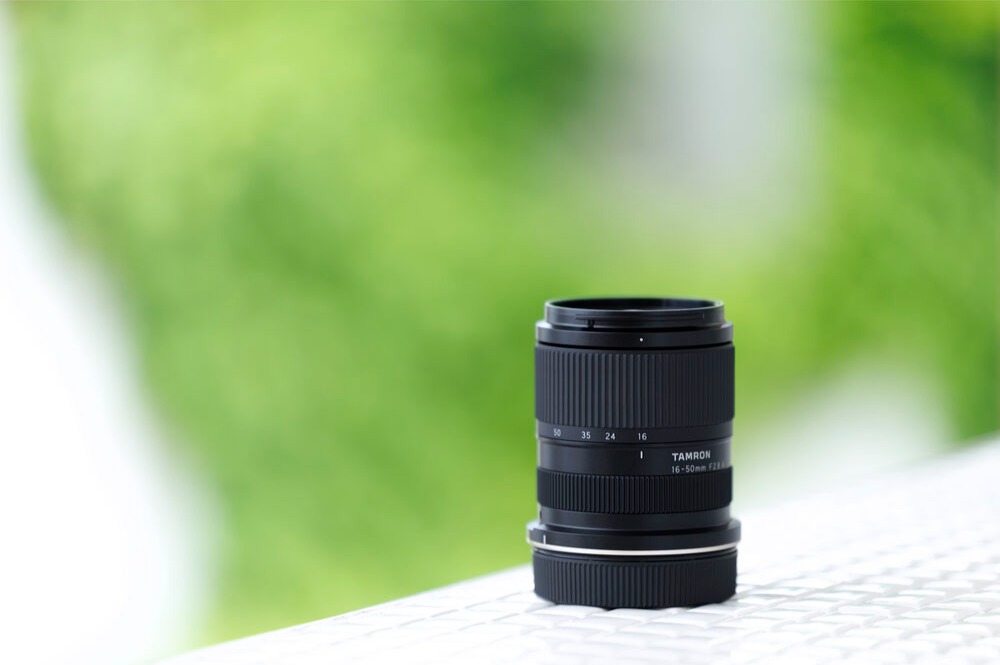 Tamron 16-50mm f/2.8 APS-C zoom lens standing on a white surface against a blurred green background.
